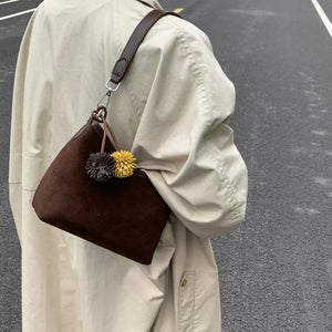 Brown suede crossbody bag with two pompom charms and a dark brown strap.
