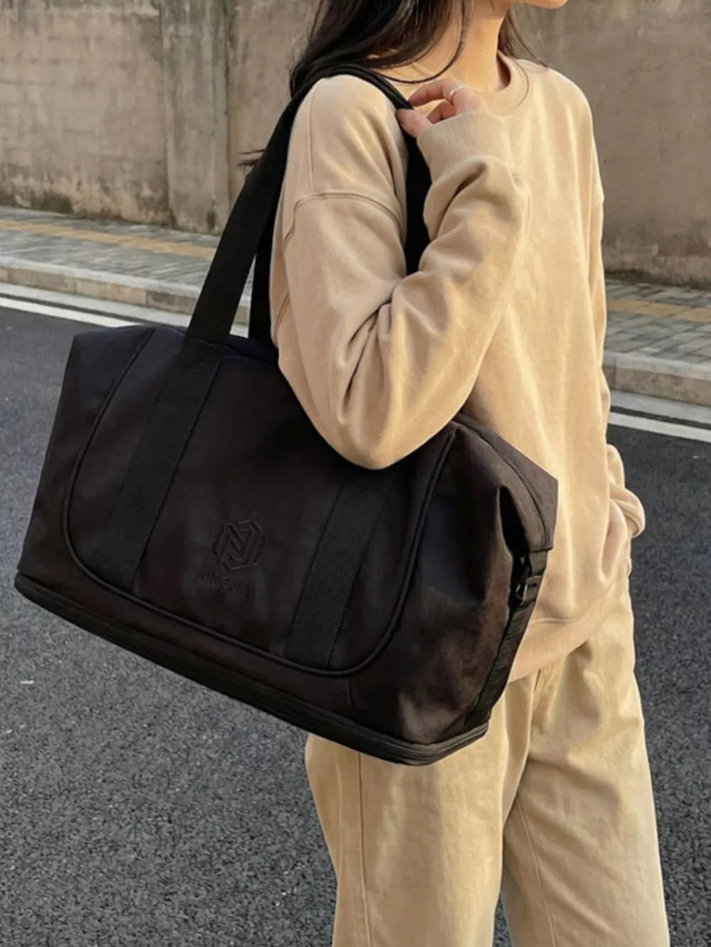 Black duffel bag with logo detail and black strap, styled with a tan sweatshirt and pants.