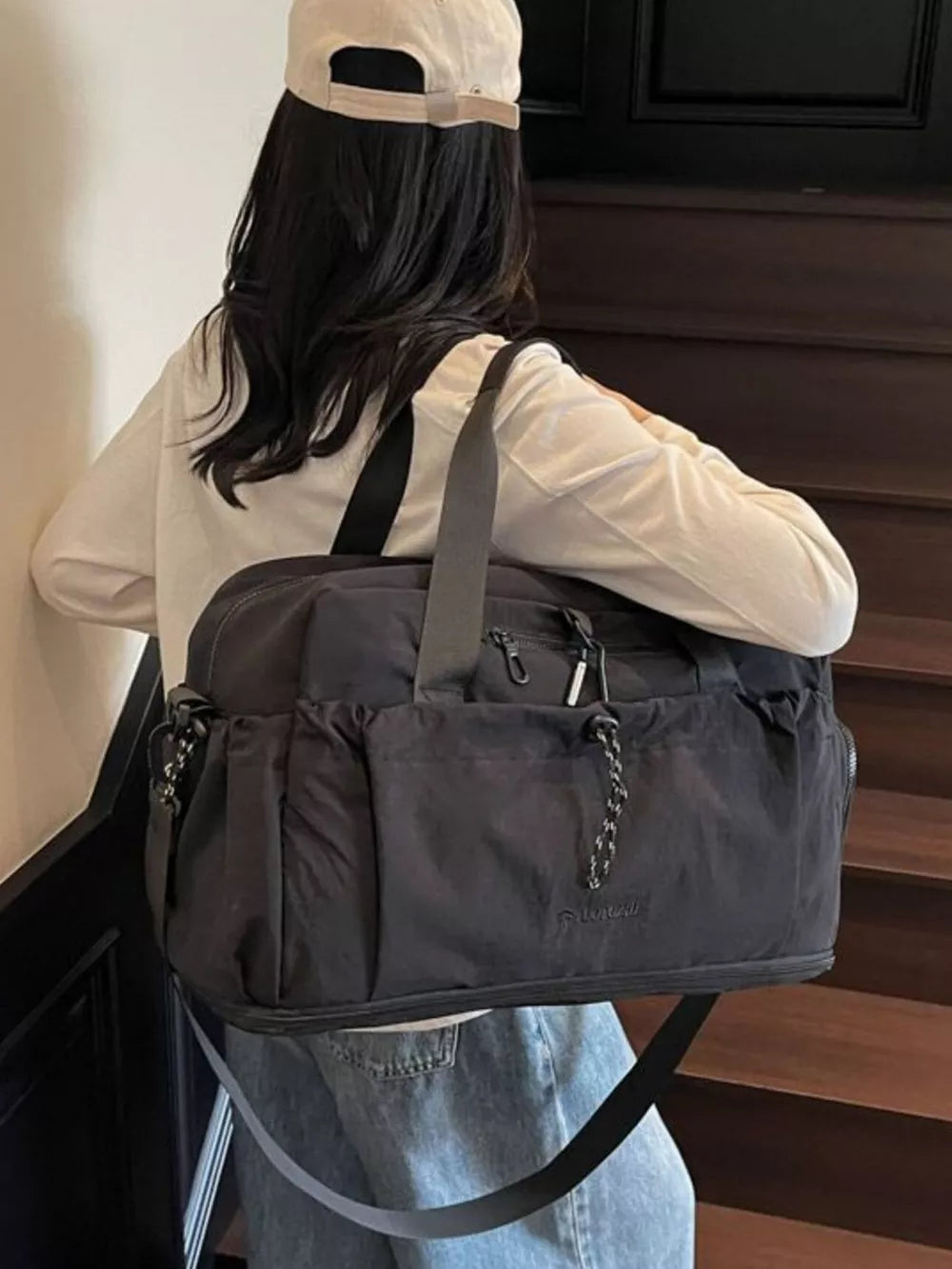 Black travel duffel bag with multiple pockets and a chain detail, worn by a person with a cap.