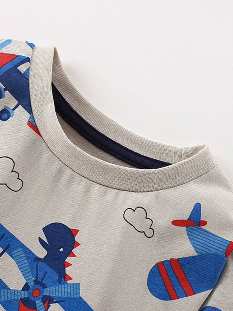 Toddler t-shirt with all-over print of blue airplanes, dinosaurs, and clouds on a light gray background.