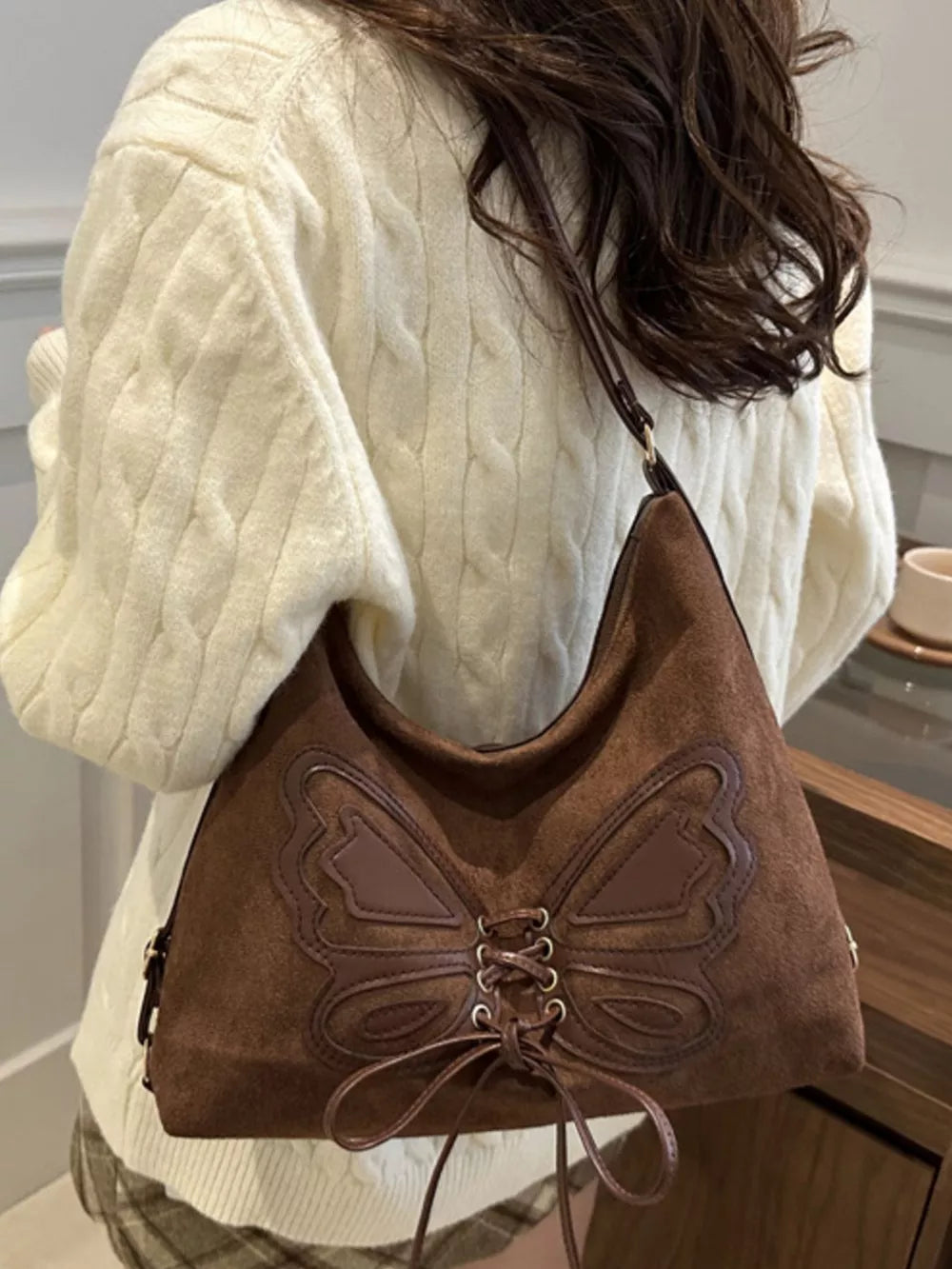 Brown suede half-moon shoulder bag with butterfly embellishment and lace-up detail.