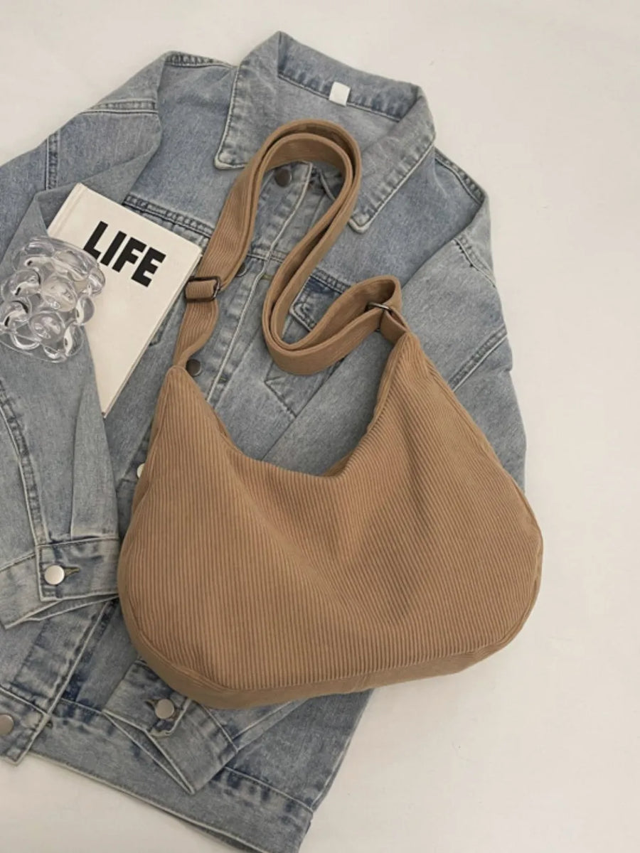 Beige corduroy crescent shoulder bag with adjustable strap and tassels, styled with denim jacket.