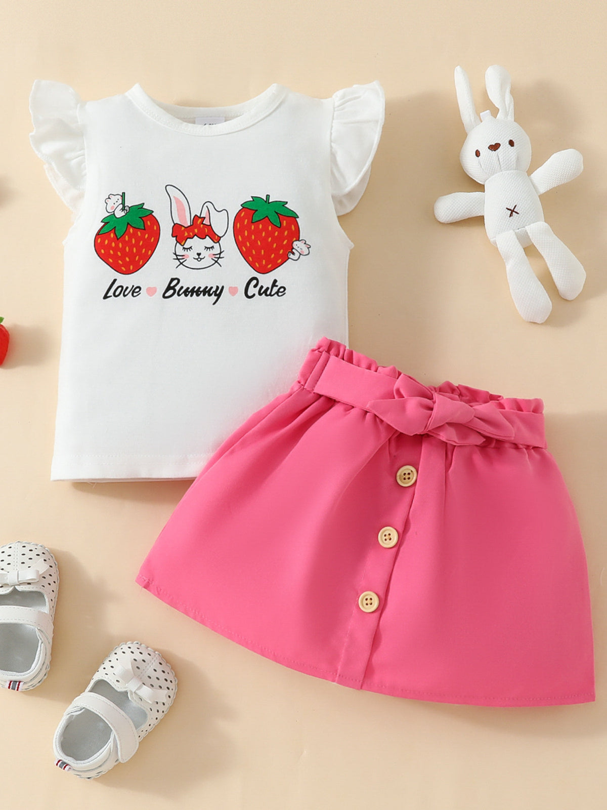 White baby set with ruffled sleeves, bunny and strawberry graphic, and pink buttoned skirt with bow detail.