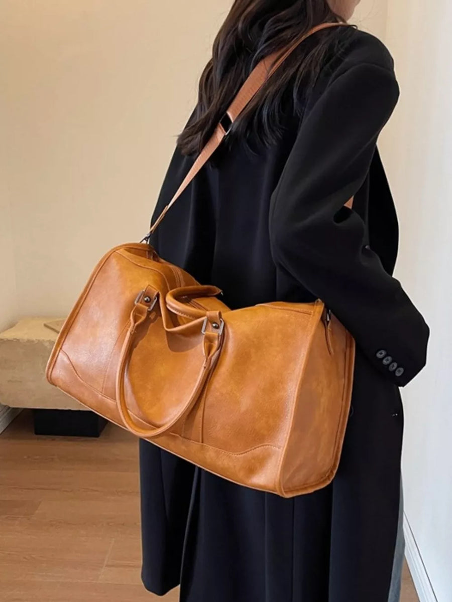 Cognac vintage leather duffle bag with top handles and shoulder strap, worn by a woman in a black coat.