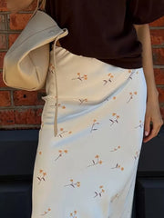 White satin midi skirt with a delicate yellow floral print and a cream shoulder bag.
