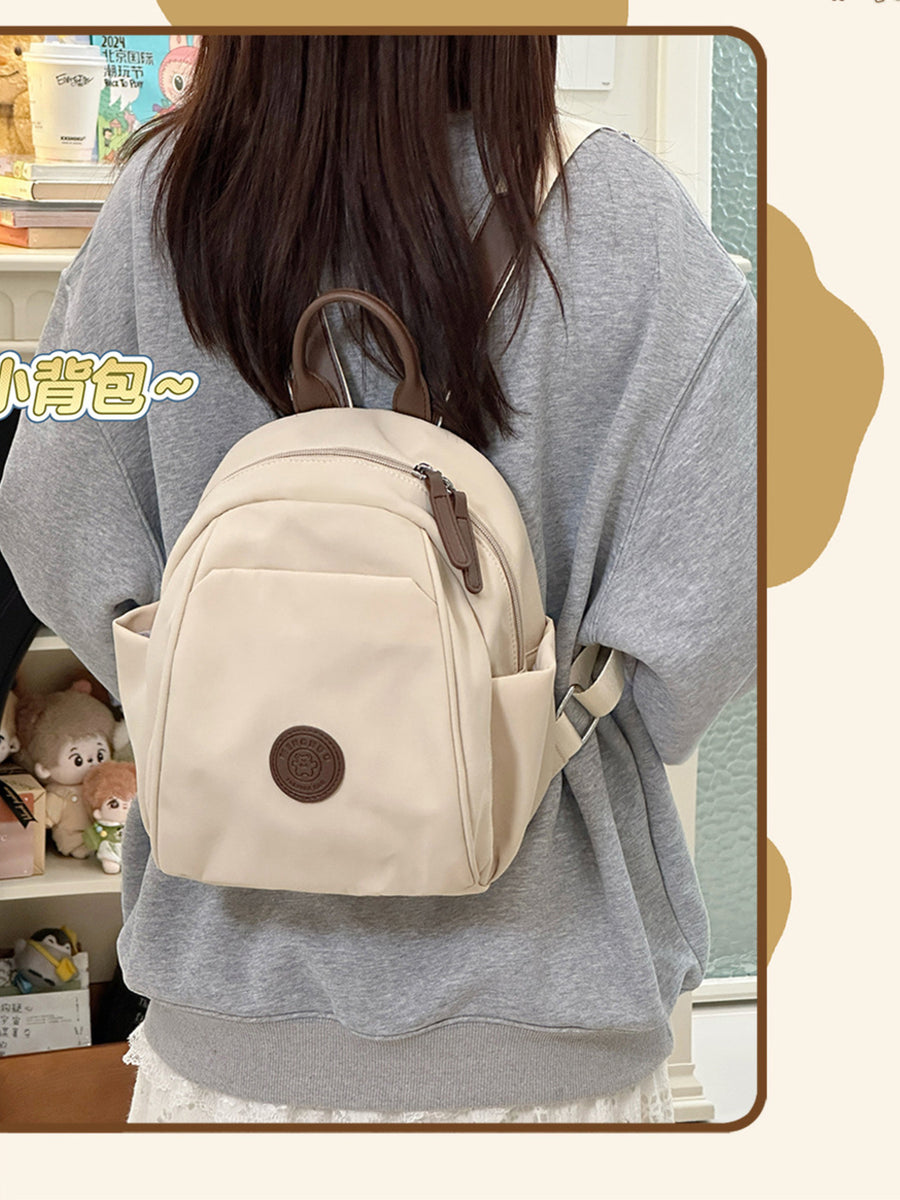 Cream mini nylon backpack with zippered pockets and bear patch detail.