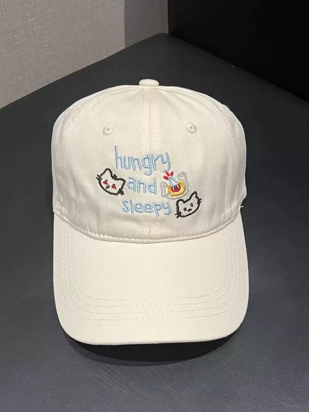 Beige baseball cap with embroidered cats and text saying hungry and sleepy