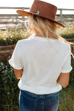 White graphic tee with rolled sleeves, styled with jeans and a brown hat.
