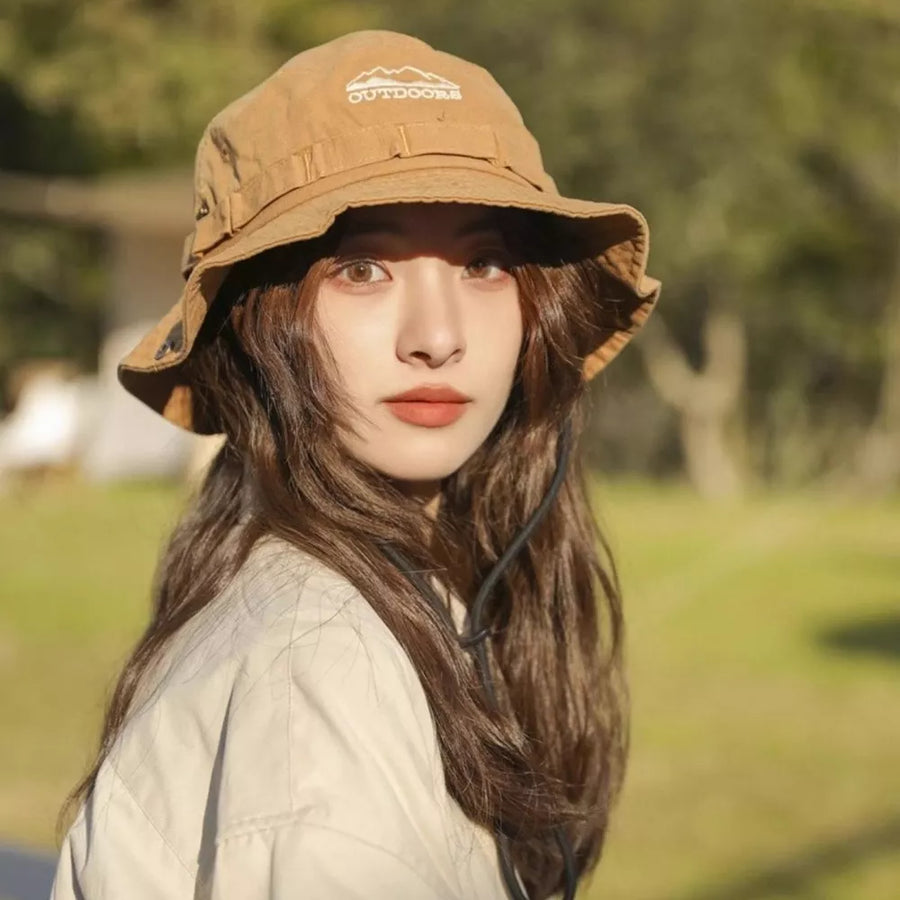 Bucket hat in tan cotton with embroidered logo and chin strap, worn with a cream shirt.