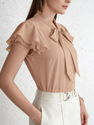 Tan tie-neck blouse with ruffle sleeves and lace trim, styled with white pants.