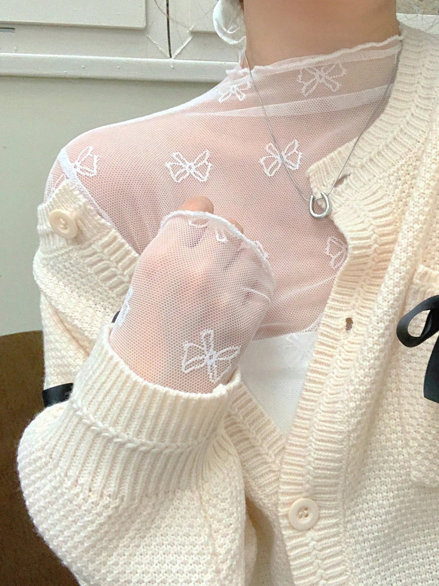 Sheer white mesh shirt with bow print and lace trim, layered under a cream knit cardigan.