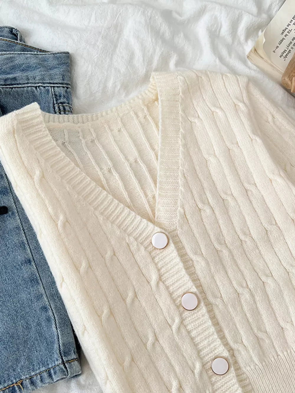 White cable-knit cropped cardigan with V-neck and button front, styled with denim.